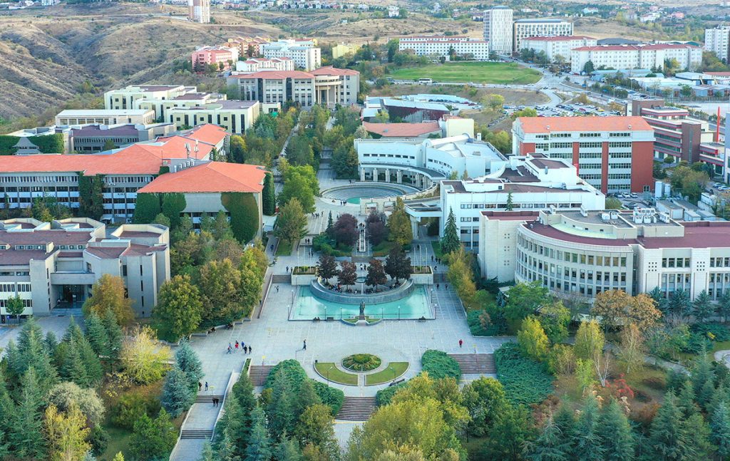 A near birds-eye-view of Bilkent University campus. An array of tall green trees and modern white buildings with red tiled roofs are arranged around a courtyard with a fountain.